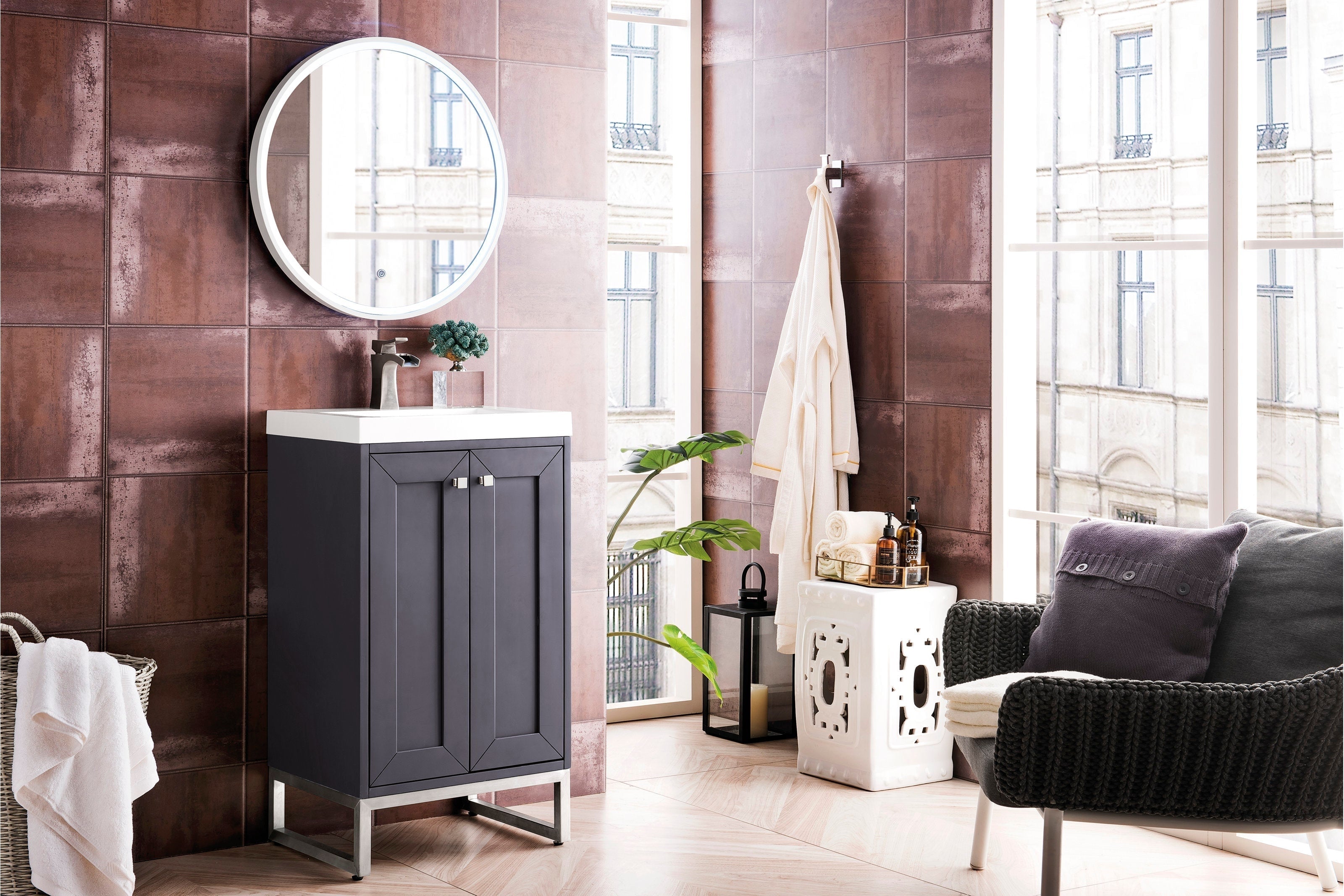 Freestanding modern solid wood bathroom vanity in a stone finish with a white stone sink top, brushed-nickel faucet and metal base, shown with a round mirror, textured brown tile wall, towel basket and decorative accessories.