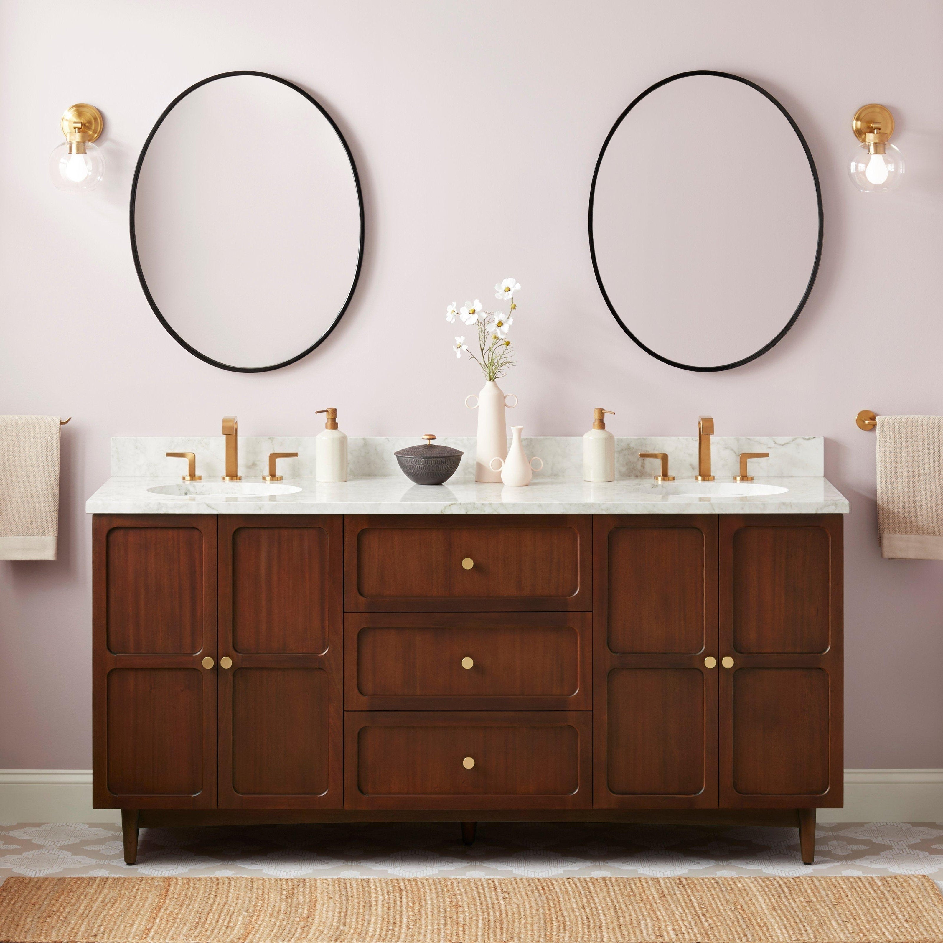 72-inch coastal solid wood double-sink vanity with Carrara marble top, two oval undermount sinks, brass faucets and knobs, round black mirrors, brass wall sconces and decorative vases against a soft pink wall.