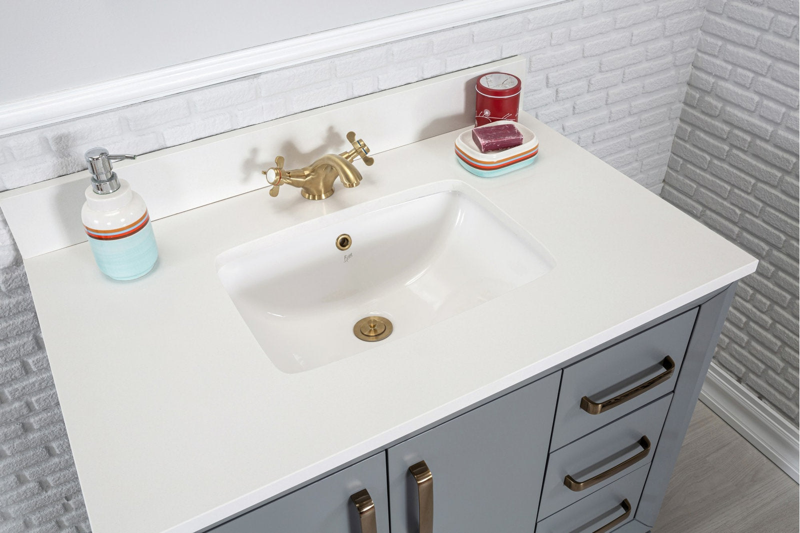 Modern gray solid-wood single-sink vanity with a white stone countertop and integrated ceramic sink, brass faucet and hardware, soap dispenser and dish on a white brick backsplash.