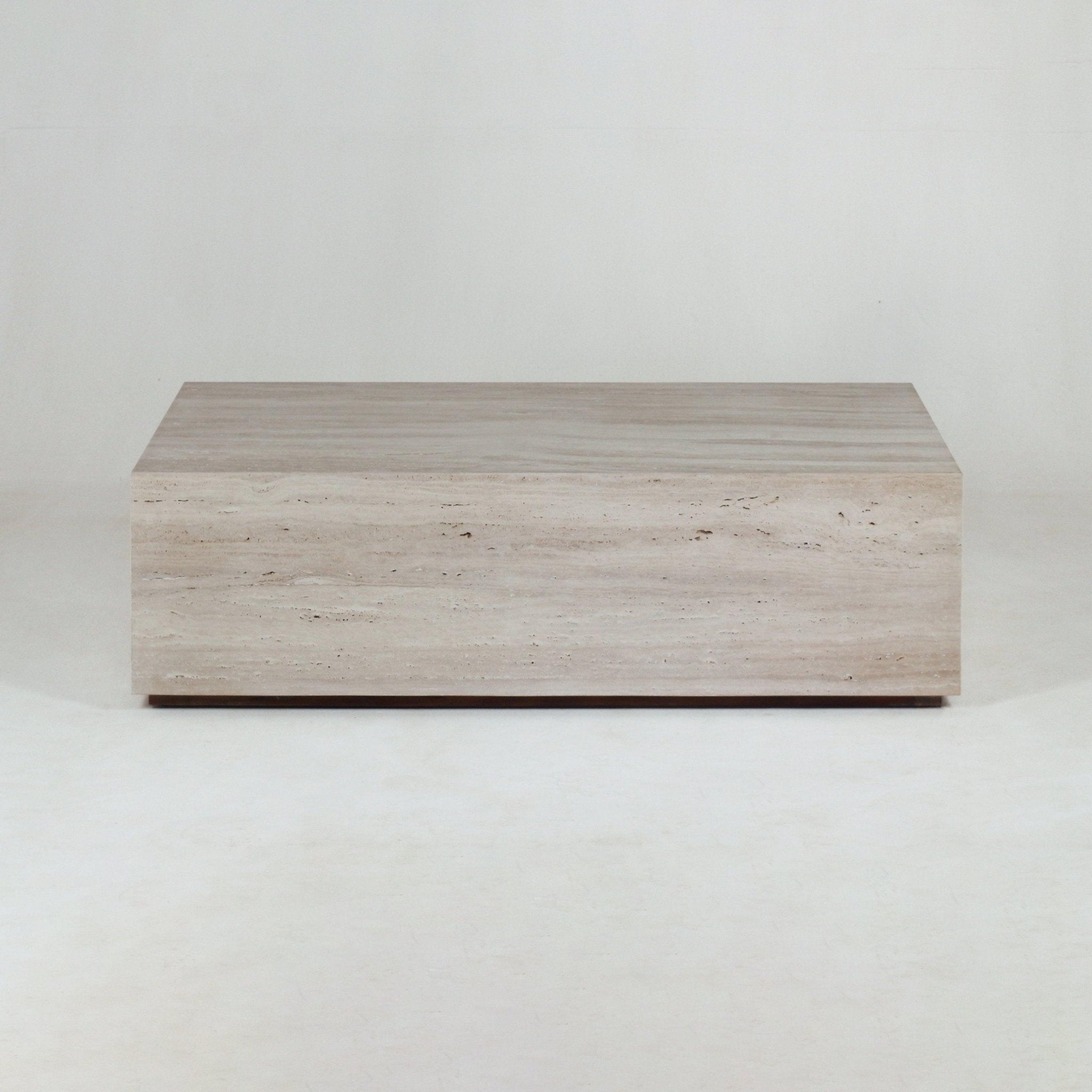 Solid rectangular travertine coffee table in natural beige with subtle horizontal veining, sitting low on a slim dark plinth against a neutral studio background.