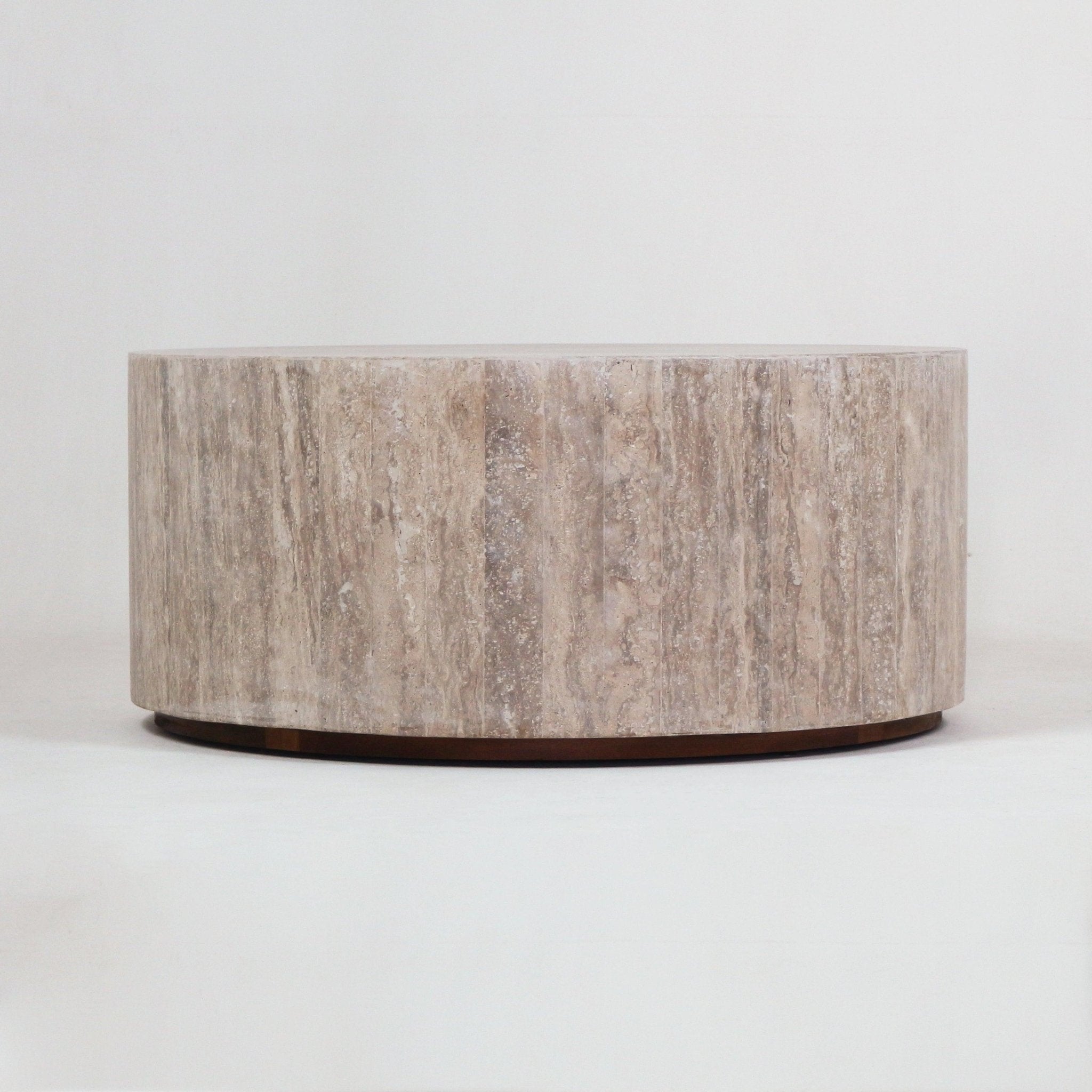 Low round coffee table carved from natural beige travertine with subtle vertical veining and a thin dark wood base, shown against a neutral background.