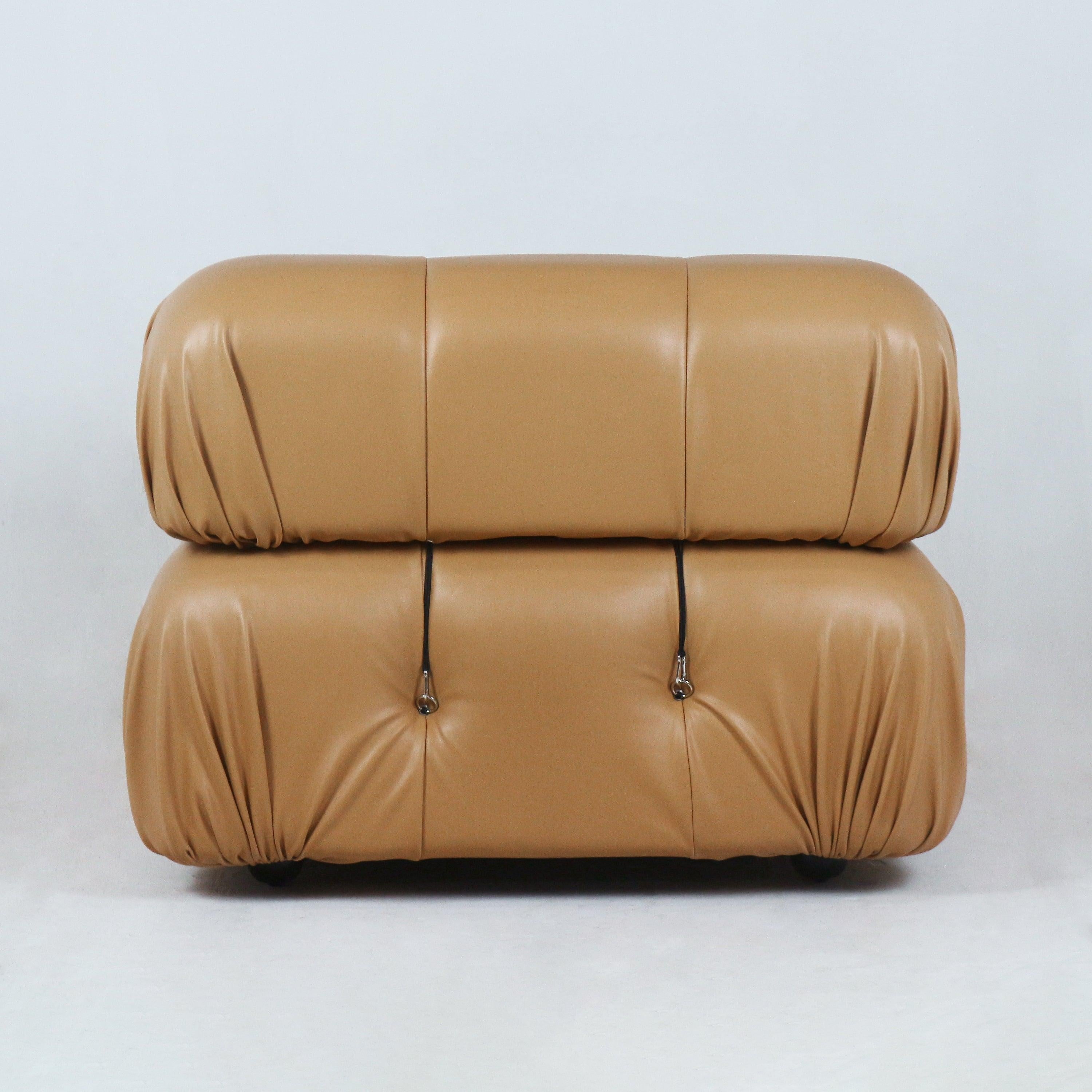 Front view of a tan leather modern sofa with two stacked rounded cushions, gathered corner pleats, tufted buttons with metal strap hardware and a low profile on short feet.