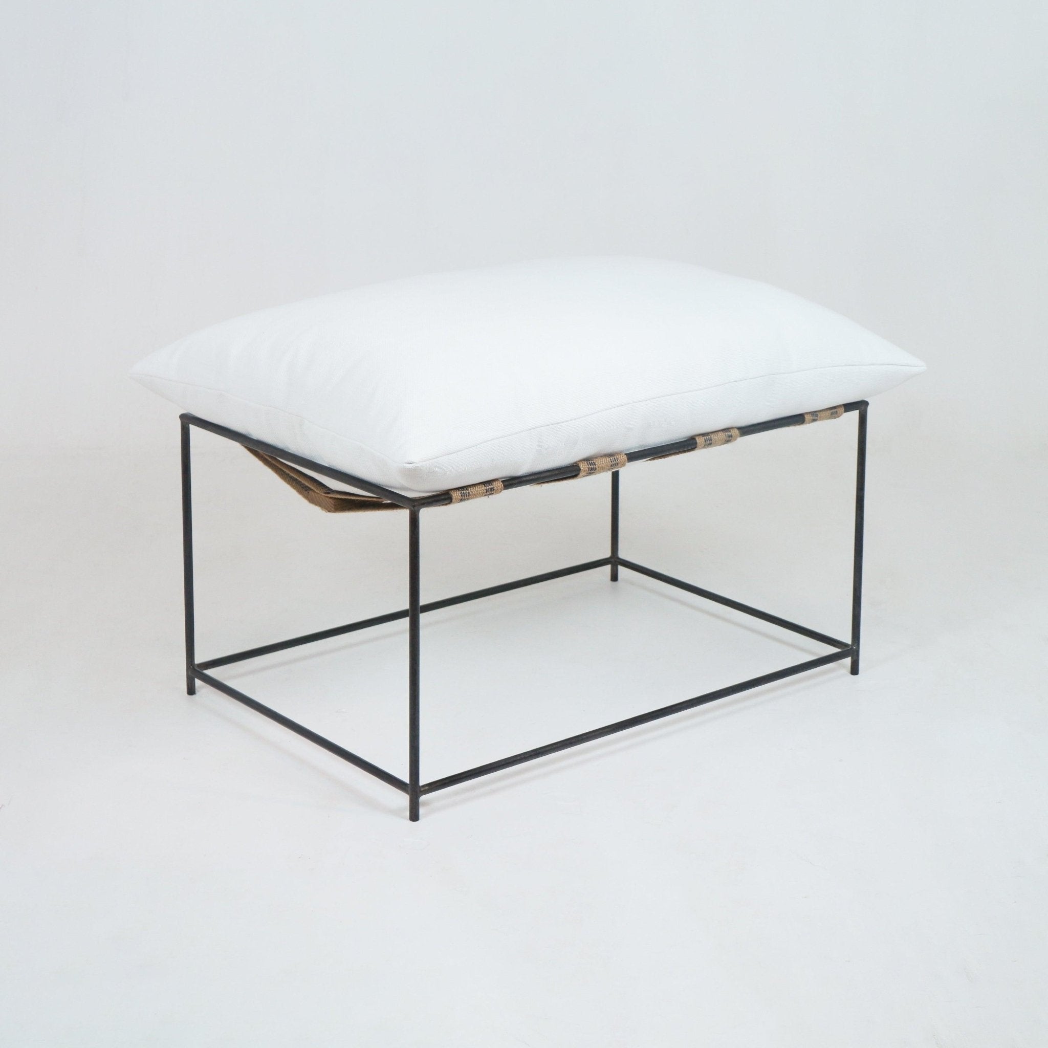 Modern ottoman featuring a plump white linen cushion atop a minimalist black metal square frame with natural woven straps, photographed on a plain white background.