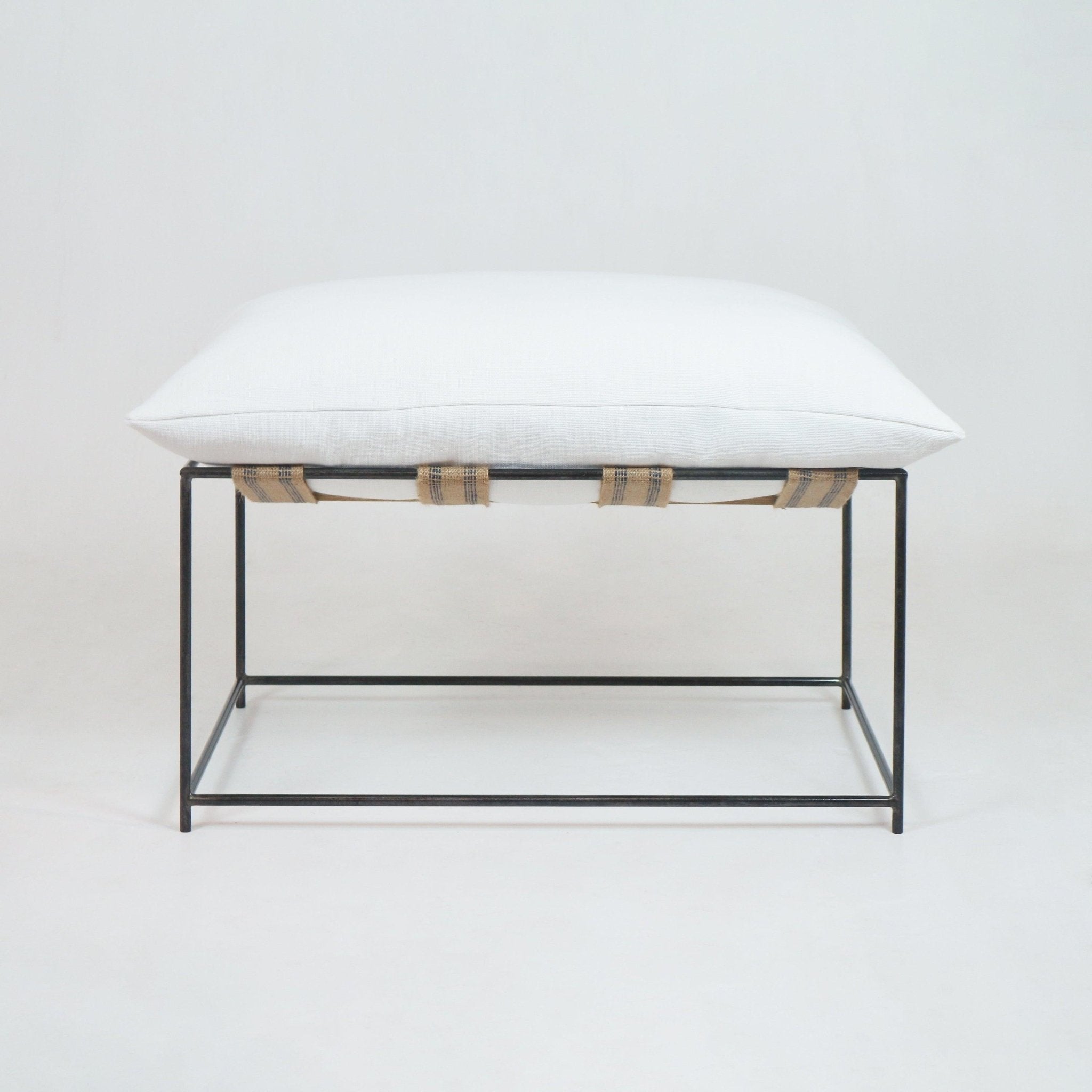 White linen cushion ottoman on a minimalist black metal frame supported by natural woven straps, photographed against a neutral background.