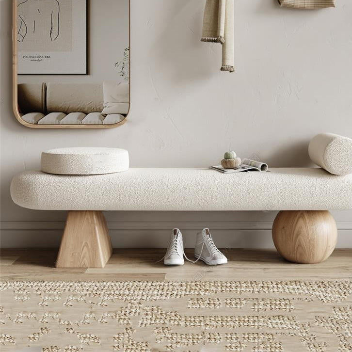 Off-white boucle upholstered backless bench with a round cushion and cylindrical bolster, supported by a natural wood pyramid and sphere leg, styled in a coastal-contemporary entryway with mirror, sneakers and textured rug.
