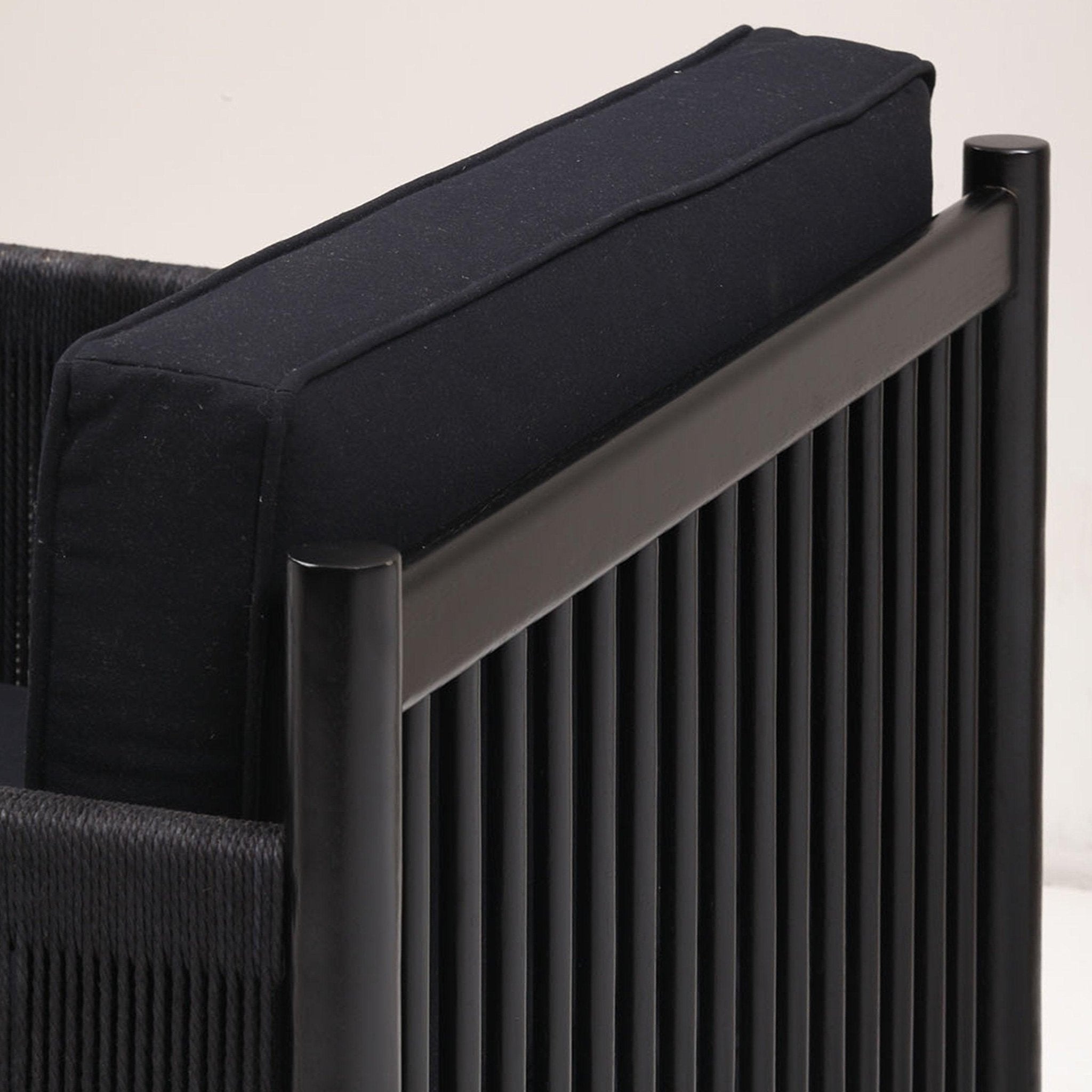 Close-up of a modern black lounge chair showing a black fabric back cushion set against a solid wood frame with rounded posts and vertical wooden spindles and woven rope arm detailing.
