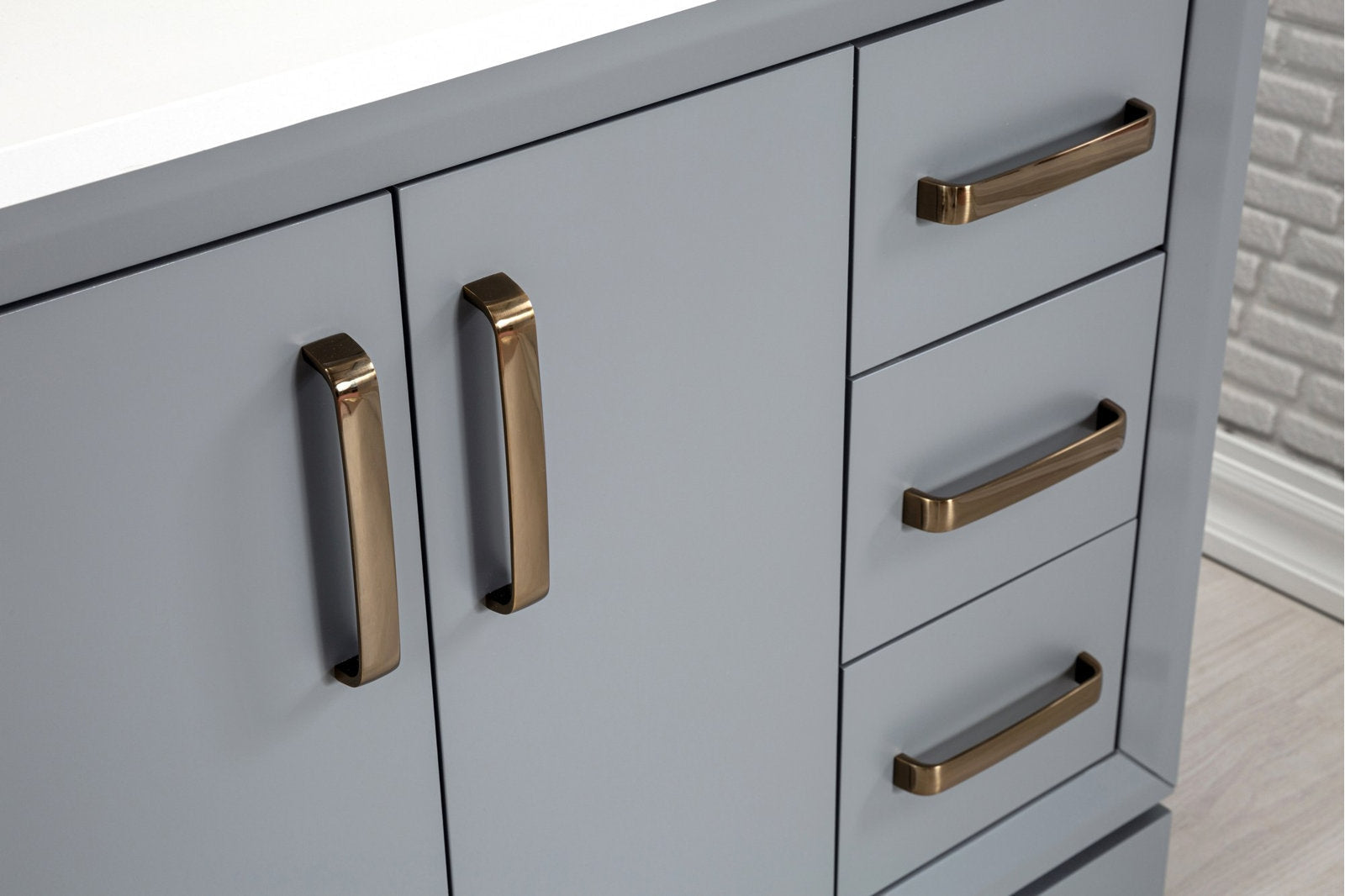Close-up of a modern stone-colored solid wood bathroom vanity showing matte gray-blue cabinet doors and stacked drawers with brushed brass handles and a white stone countertop edge.
