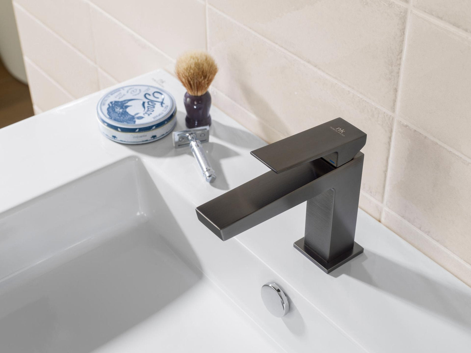 Close-up of a modern matte-black single-handle faucet mounted on a white ceramic sink with a round chrome overflow button, beige stone tile backsplash and shaving accessories (brush, tin and razor) on the counter.
