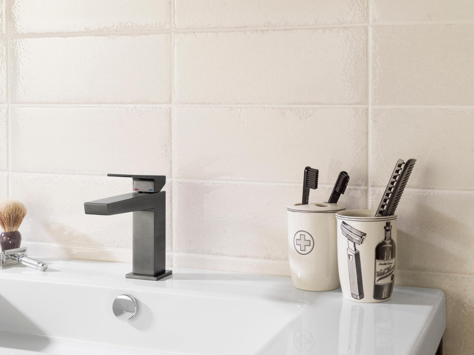 Modern single-sink vanity with a stone-colored tiled backsplash, white ceramic sink and square chrome faucet, shown with two decorative toothbrush holders and shaving accessories on the countertop.