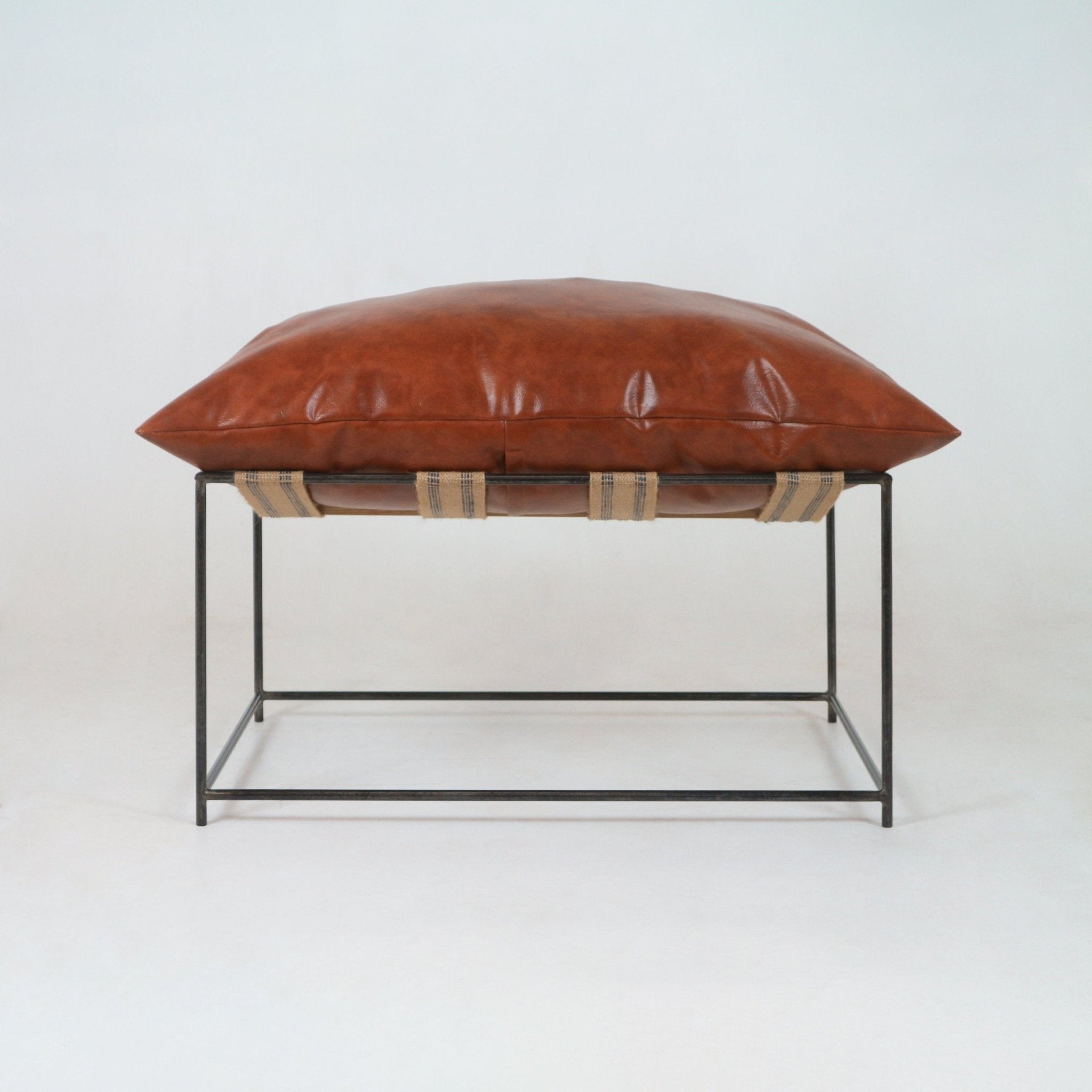 Low-profile ottoman with a plush red leather cushion resting on a minimalist black metal square frame, supported by natural woven webbing straps — a modern living room accent stool.