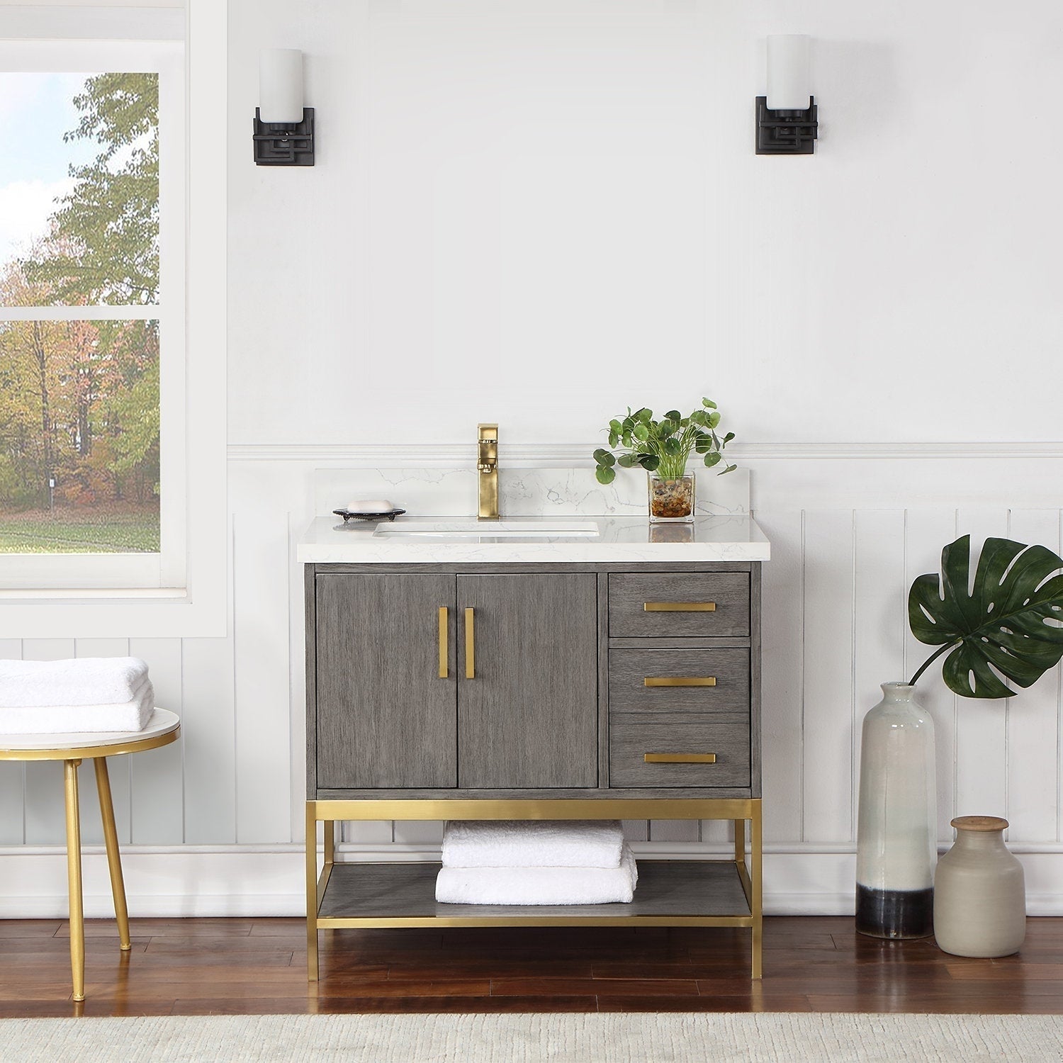 36-inch freestanding modern gray solid-wood vanity with stone countertop and white undermount sink, brass faucet, brass handles and base, two cabinet doors, three drawers and a lower shelf with folded white towels.