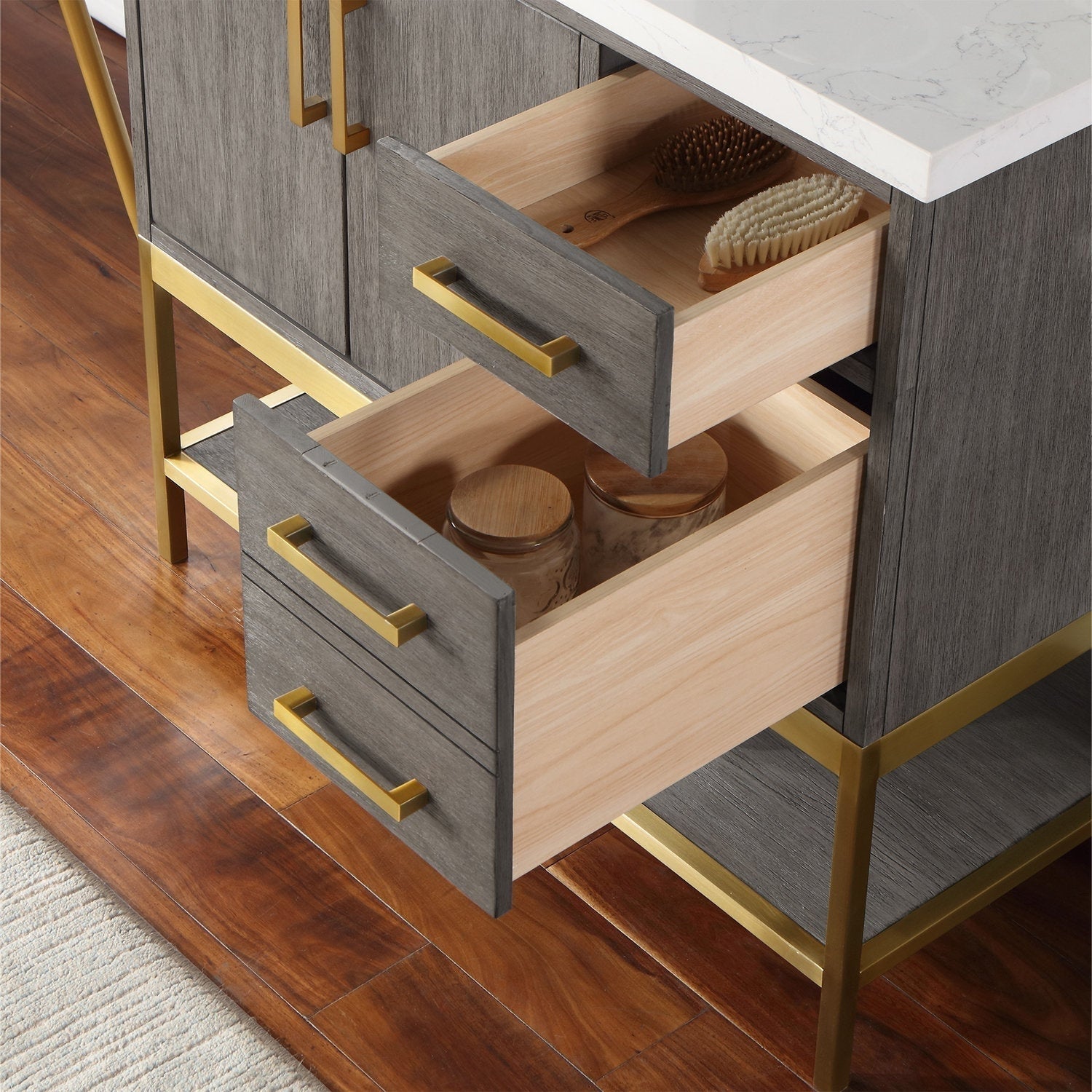 Close-up of a modern stone-top bathroom vanity showing gray wood veneer drawer fronts with brass-finish handles and metal base; two drawers open to reveal light wood interiors holding brushes and storage jars.